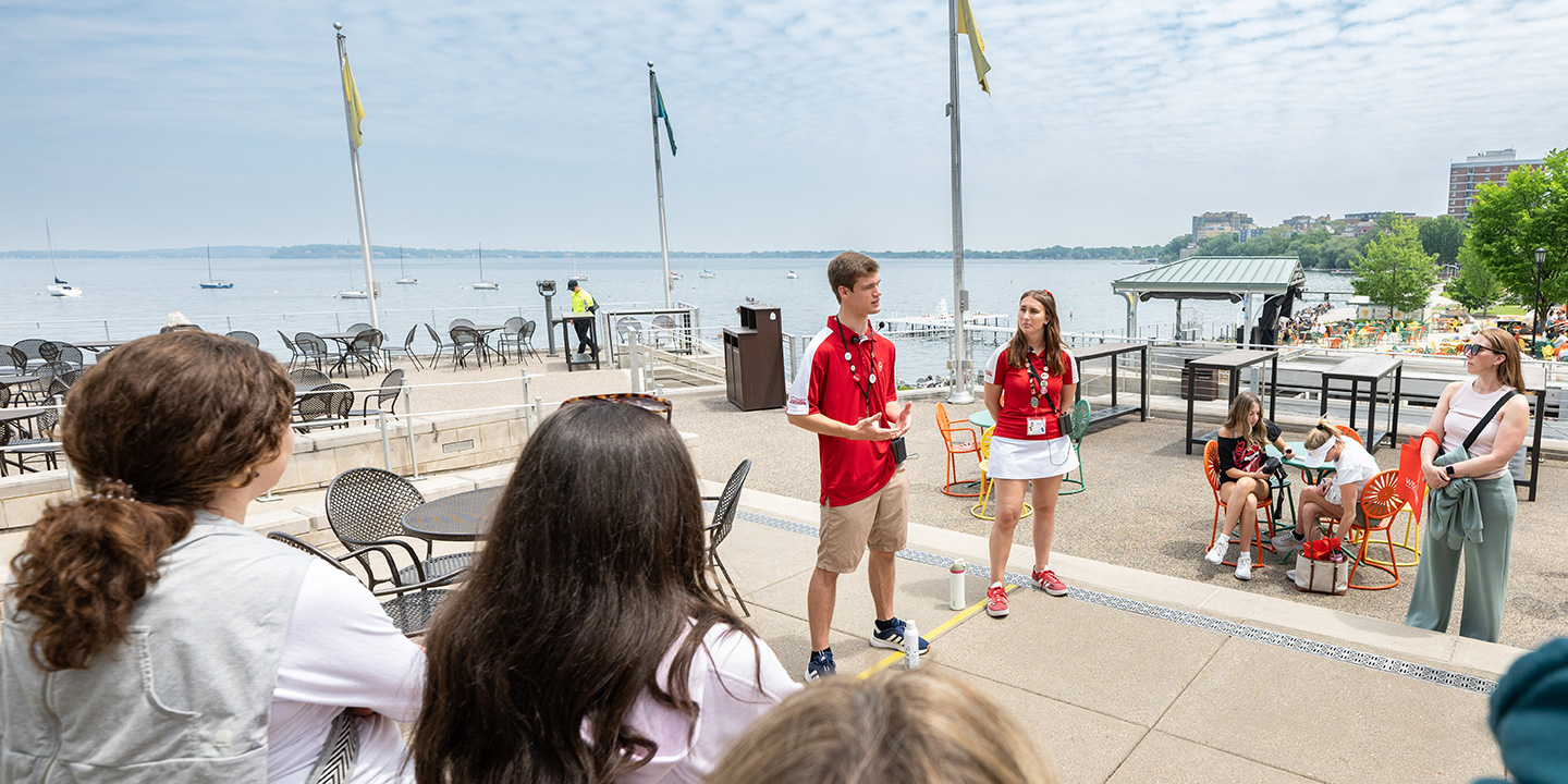 Two tour guides in red shirts speak to a small group on a lakeside terrace with colorful chairs, flags, and boats visible on the water under a partly cloudy sky.