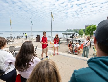 Two tour guides in red shirts speak to a small group on a lakeside terrace with colorful chairs, flags, and boats visible on the water under a partly cloudy sky.