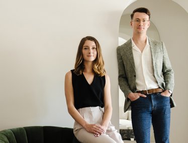 Grace and Michael van Meurer are posed indoors in a modern, minimalist setting, with one seated on a dark sofa and the other standing near an arched doorway.