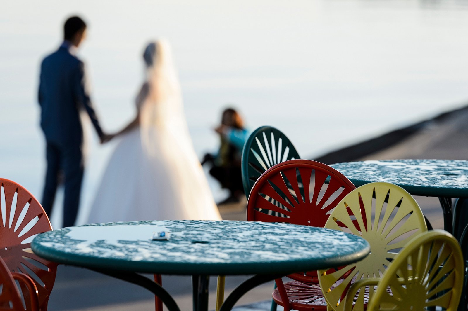 Colorful terrace chairs and tables in the foreground with a blurred bride and groom holding hands with Lake Mendota in the background.