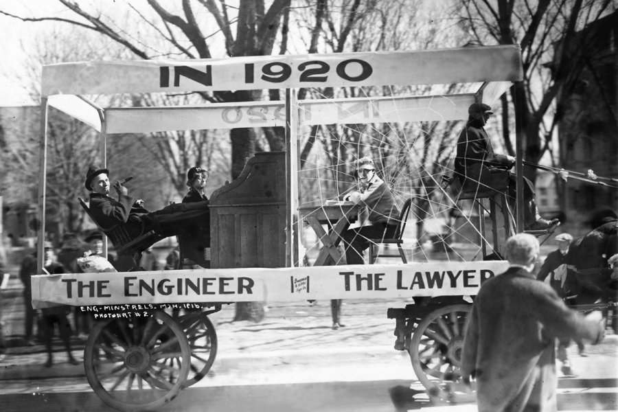 A black‑and‑white photo of a parade float built on a wagon with large wooden wheels. The float has a canopy reading 'IN 1920' and features two seated participants, one positioned under a sign labeled 'The Engineer' and the other under 'The Lawyer.' The float is surrounded by onlookers, and bare trees indicate a chilly season.