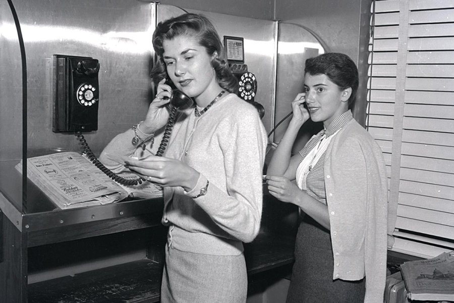 A black‑and‑white photograph of two women standing at adjacent public telephone booths. Each is holding a rotary handset while referencing a phone directory.