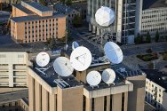 An aerial view shows the rooftop of the Atmospheric, Oceanic and Space Sciences Building, covered with multiple large white satellite dishes and antennas, surrounded by nearby offices and streets.