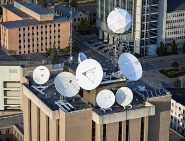 An aerial view shows the rooftop of the Atmospheric, Oceanic and Space Sciences Building, covered with multiple large white satellite dishes and antennas, surrounded by nearby offices and streets.
