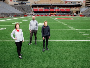 Three people wearing Wisconsin athletic wear stand spaced apart on the turf of Camp Randall on the otherwise empty field.