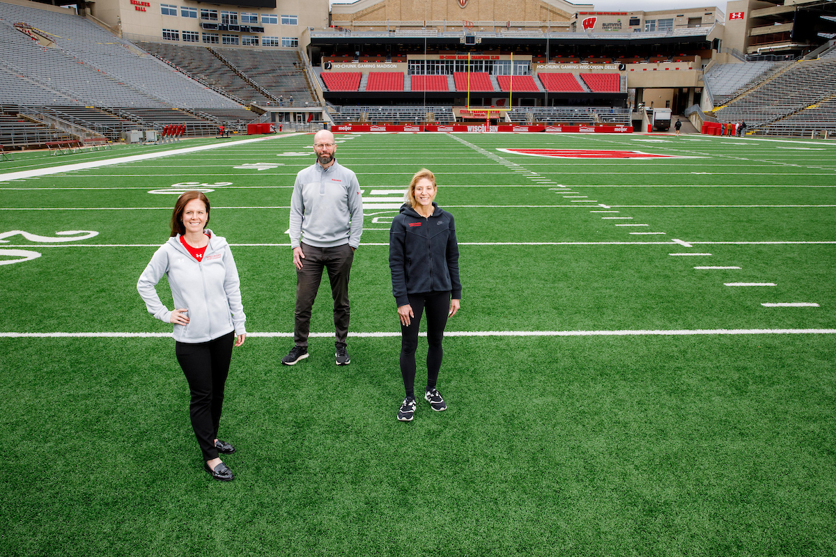 Three people wearing Wisconsin athletic wear stand spaced apart on the turf of Camp Randall on the otherwise empty field.