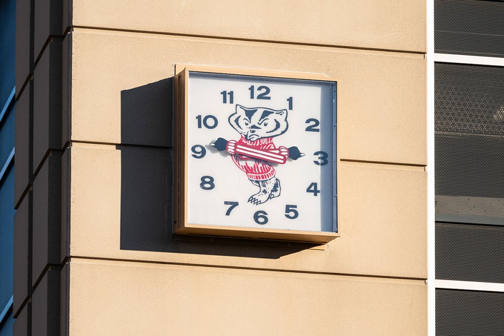 A square outdoor clock mounted on a building features Bucky Badger in his striped red and white sweater at its center with his arms acting as the hands of the clock.