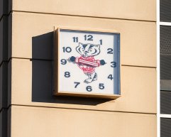 A square outdoor clock mounted on a building features Bucky Badger in his striped red and white sweater at its center with his arms acting as the hands of the clock.