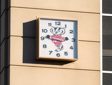 A square outdoor clock mounted on a building features Bucky Badger in his striped red and white sweater at its center with his arms acting as the hands of the clock.