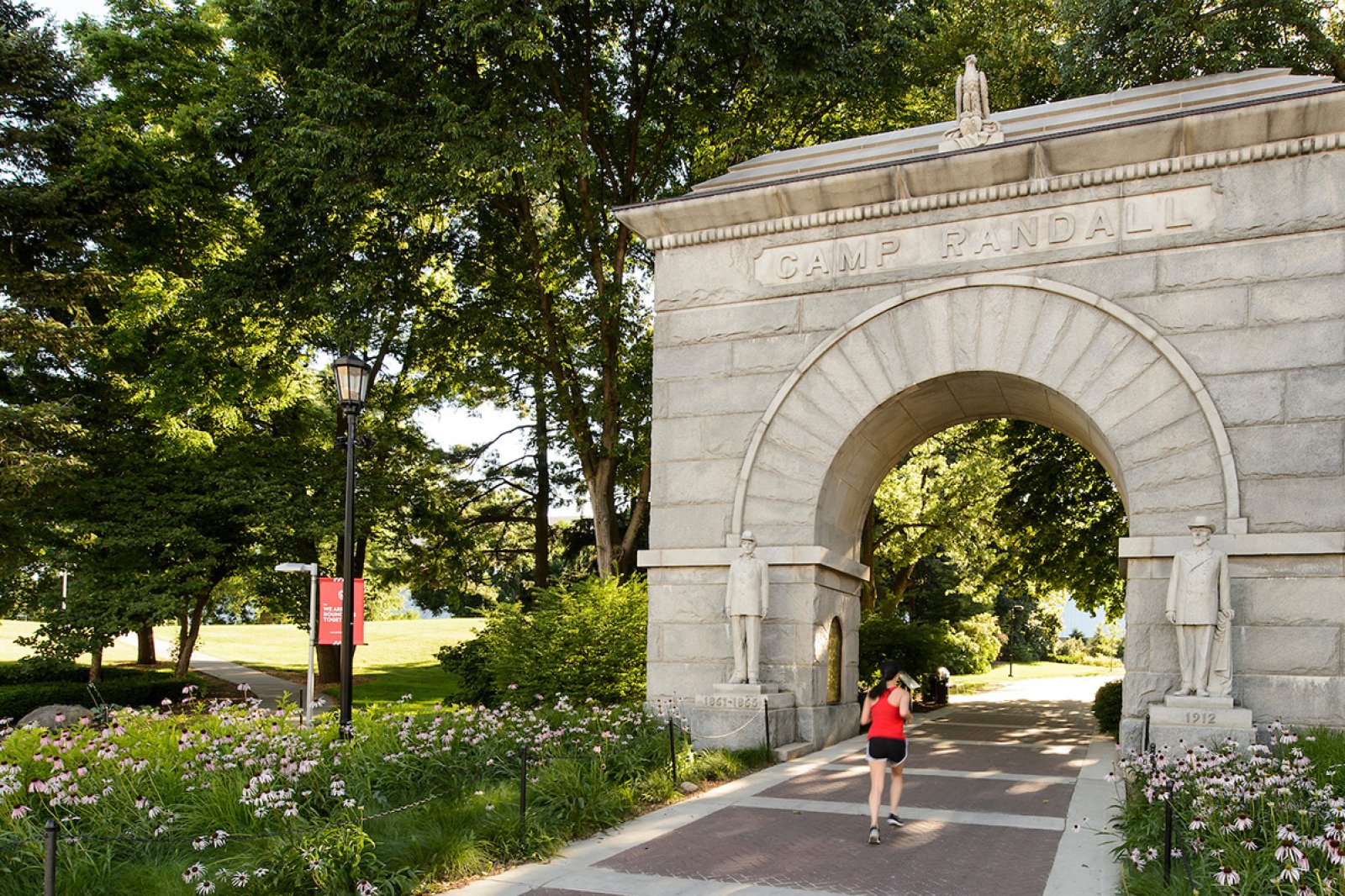 A person jogs along a flower-lined brick path that goes through the large stone Camp Randall Memorial Arch, surrounded by trees and greenery.