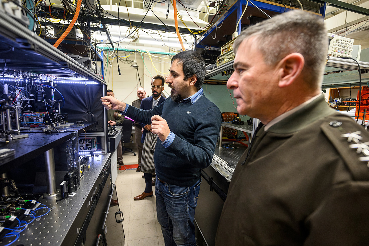A group of people, one in military dress, tour a laboratory filled with complex electronic equipment and wiring as one person gestures toward an illuminated setup.