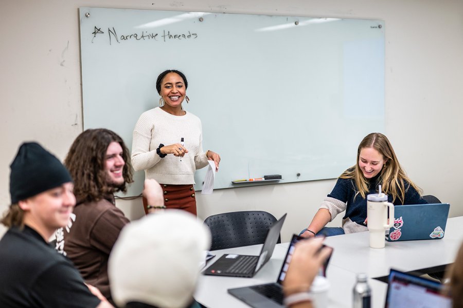 Dantiel Moniz stands at the front of a classroom leading a discussion while several students sit at a table using laptops and taking notes.