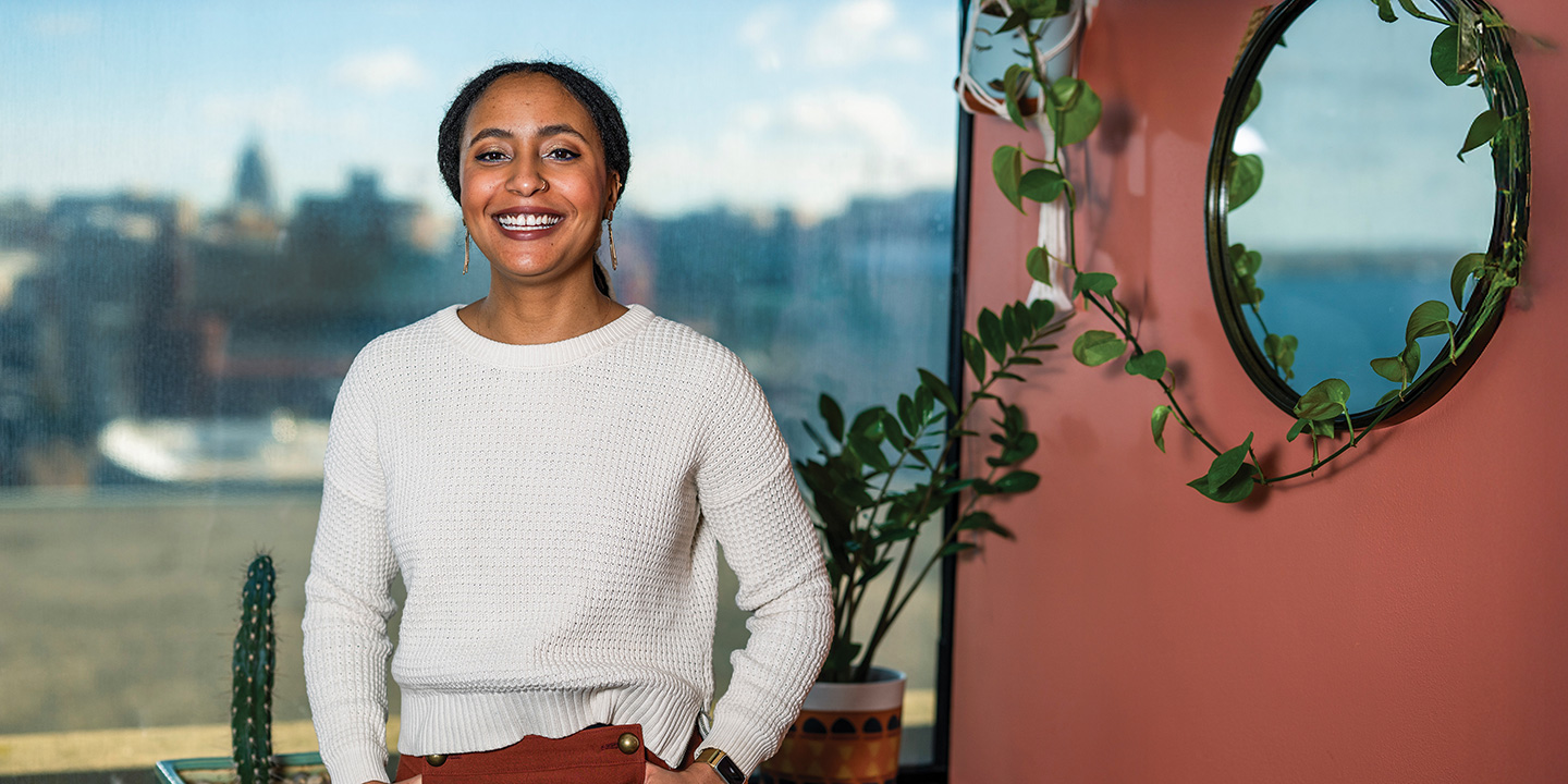 Dantiel Moniz stands indoors near a window with a cityscape in the background and a leafy plant hanging beside a round mirror on the wall.