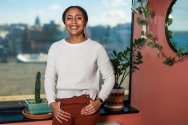 Dantiel Moniz stands indoors near a window with a cityscape in the background and a leafy plant hanging beside a round mirror on the wall.