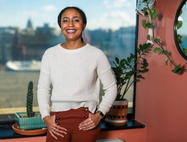 Dantiel Moniz stands indoors near a window with a cityscape in the background and a leafy plant hanging beside a round mirror on the wall.