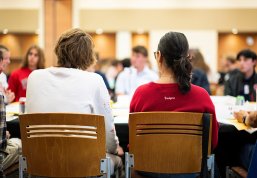 People sit at a round table in a bright, busy meeting room, participating in a group discussion.