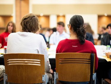 People sit at a round table in a bright, busy meeting room, participating in a group discussion.