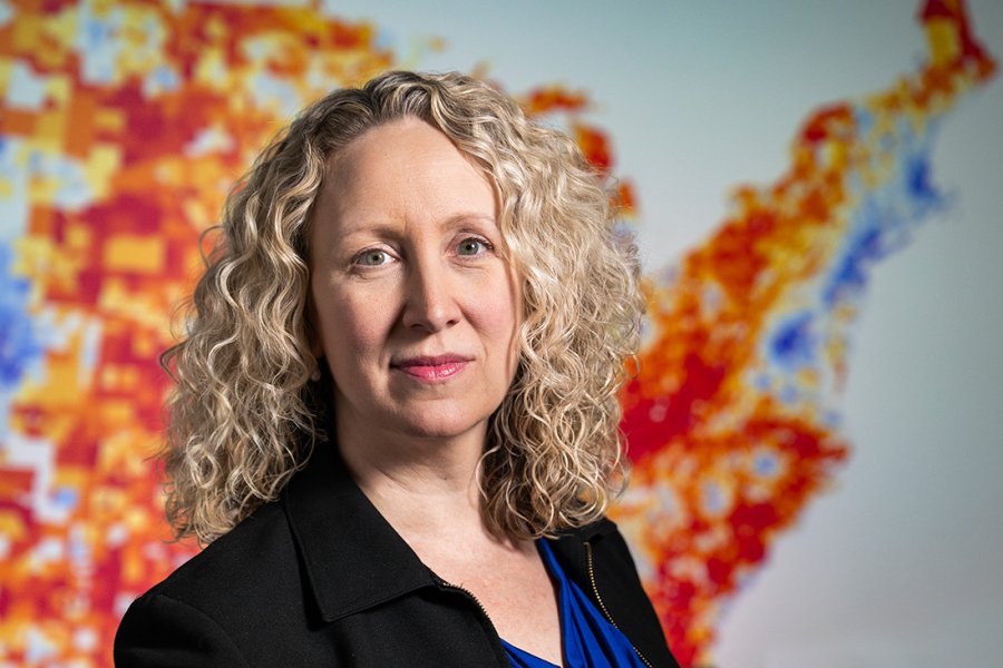Dr. Amy Kind, a woman with curly blonde hair stands in front of the colorful ADI map of Wisconsin that uses red, orange, yellow, and blue shading to represent varying levels of community disadvantage.