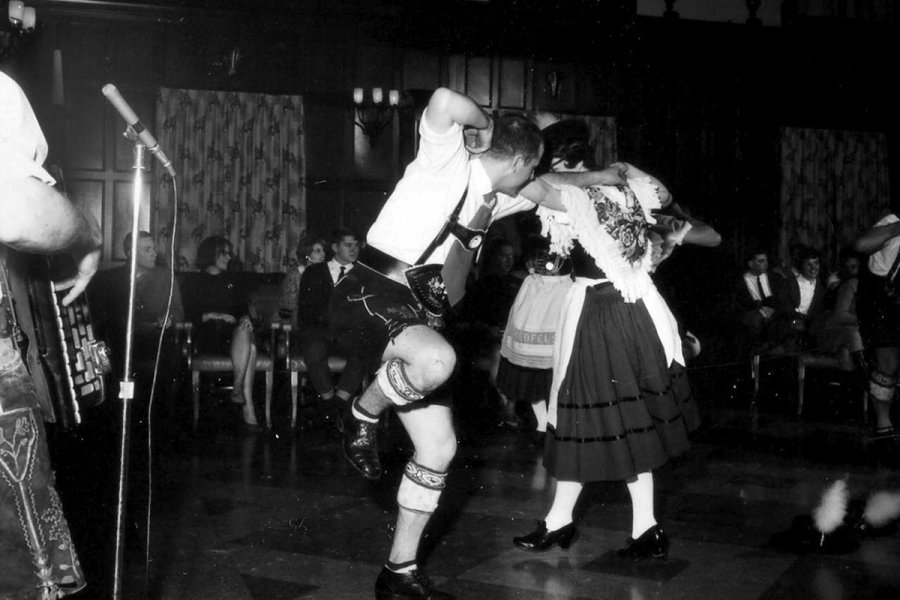 Black‑and‑white photograph of a man and woman dancing indoors, with the man wearing traditional lederhosen and the woman wearing a full skirt and embroidered shawl. Other performers and seated spectators are visible in the background.