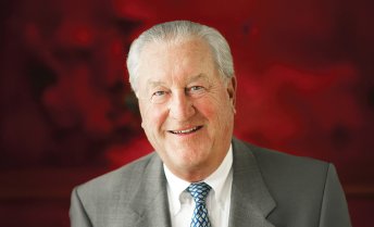 Portrait of Jerry Frautschi wearing a gray suit jacket, white dress shirt, and patterned blue tie, in front of a deep red background.
