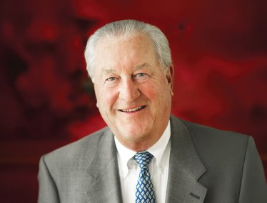 Portrait of Jerry Frautschi wearing a gray suit jacket, white dress shirt, and patterned blue tie, in front of a deep red background.