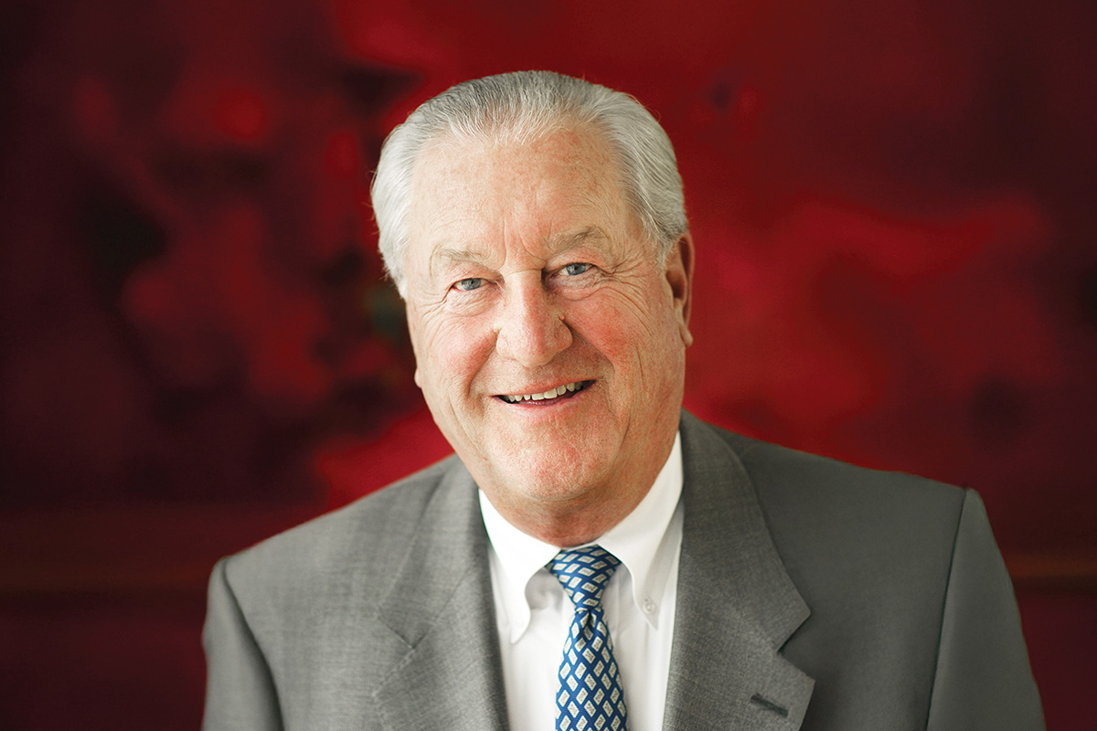 Portrait of Jerry Frautschi wearing a gray suit jacket, white dress shirt, and patterned blue tie, in front of a deep red background.