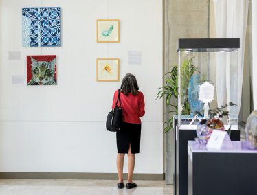 A person stands in front of a gallery wall observing four colorful framed artworks, with additional glass and ceramic sculptures displayed in cases nearby.