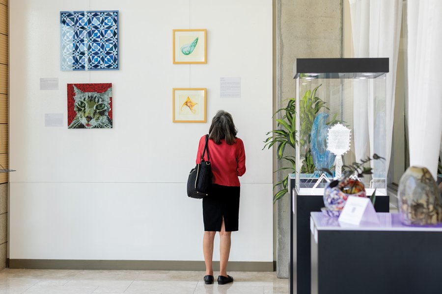 A person stands in front of a gallery wall observing four colorful framed artworks, with additional glass and ceramic sculptures displayed in cases nearby.