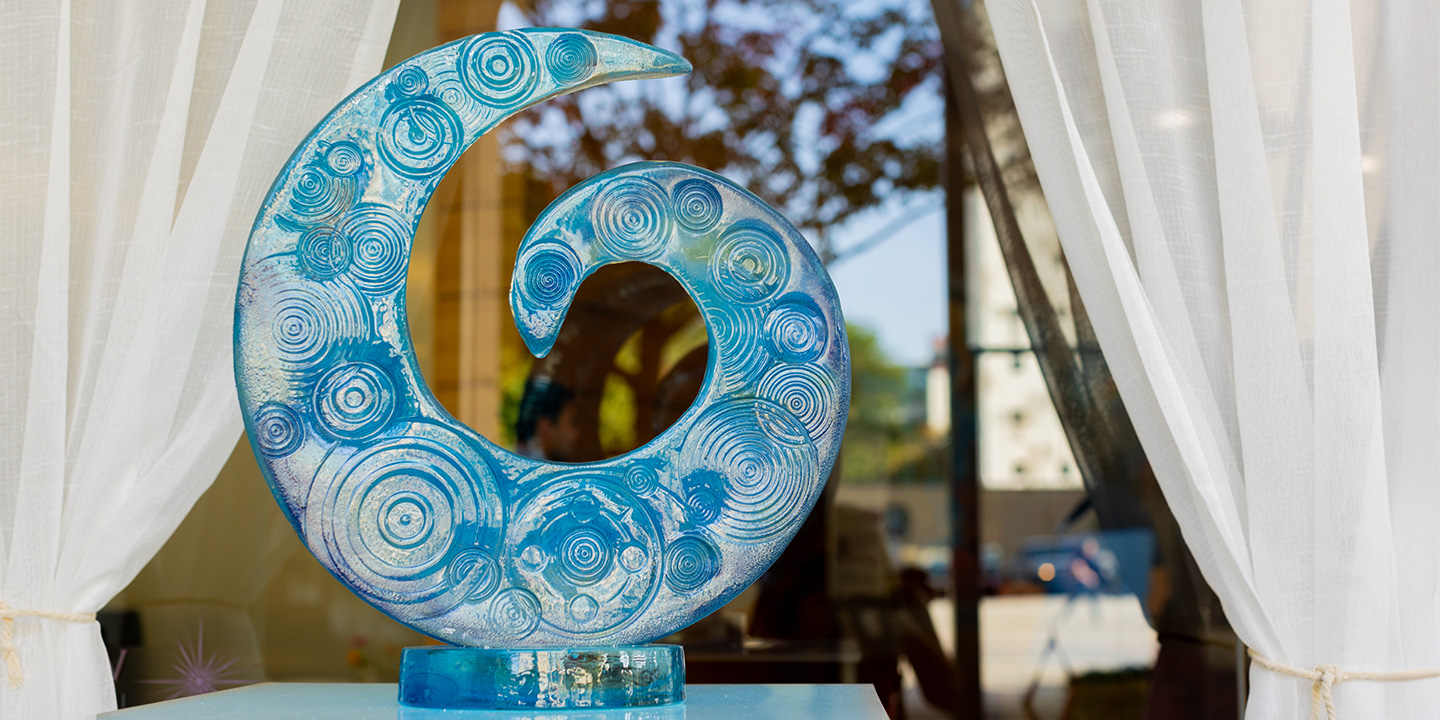 A blue glass sculpture with circular patterns, shaped like a curling wave or spiral, is displayed on a pedestal between white curtains.