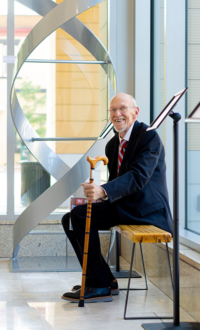 John Wiley, wearing a suit, sits on a small bench holding a wooden cane near a large metal helix sculpture inside a bright gallery space.