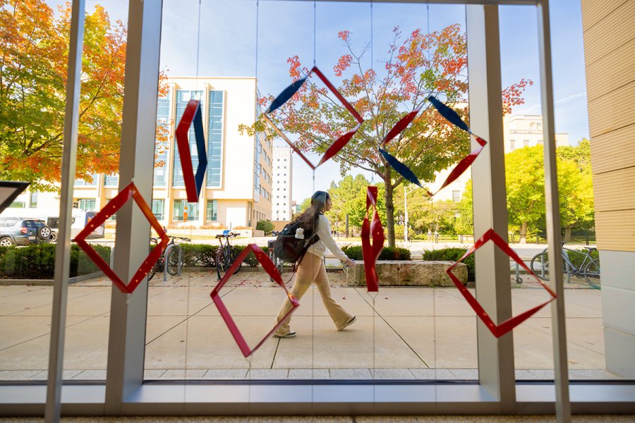 A person walks outside past large gallery windows decorated with hanging red and blue geometric shapes, with autumn trees and campus buildings in the background.