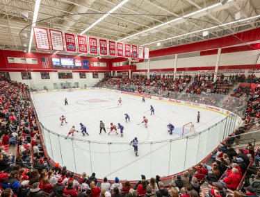 A packed arena watches as two hockey teams face off at center ice inside a brightly lit rink surrounded by cheering fans.