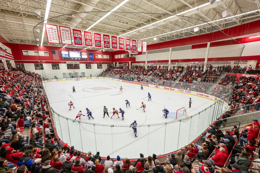 A packed arena watches as two hockey teams face off at center ice inside a brightly lit rink surrounded by cheering fans.