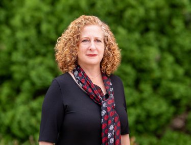 Chancellor Mnookin poses outdoors in front of a lush green background, wearing a dark top and a patterned scarf.