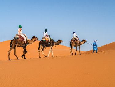 A line of three people ride camels across a bright orange sand dune while a guide in blue clothing leads them through the desert under a clear blue sky.