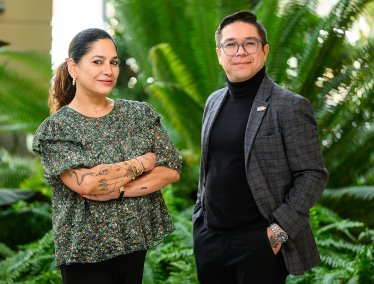 A male and female stand side by side in a lush outdoor setting with large green plants behind them, the female wearing a patterned top with visible tattoos and the male dressed in a dark turtleneck and blazer.