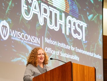 Chancellor Mnookin stands at a wooden podium in front of a large screen displaying the ‘Earth Fest’ logo .