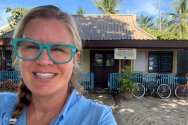 Sarah Spelsberg, wearing a blue shirt, stands outdoors in a sandy area with palm trees in the background.