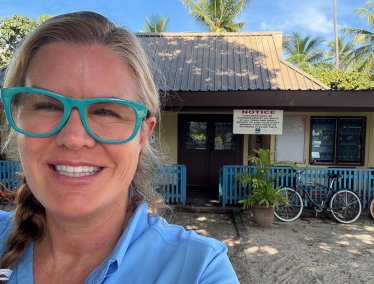 Sarah Spelsberg, wearing a blue shirt, stands outdoors in a sandy area with palm trees in the background.