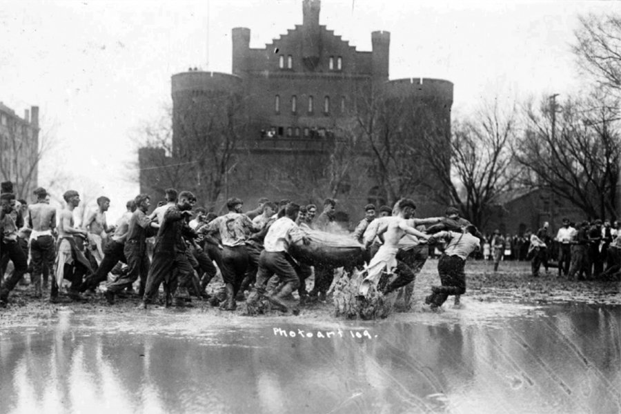A historic black‑and‑white photograph of a group of young men engaged in a muddy game with a large sack in rain‑soaked field Library Mall. The Red Gym is in the background.