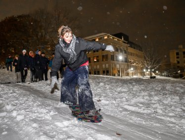 A female, smiling, snowboards down a snowy Bascom Hill at night while a group of people watch.