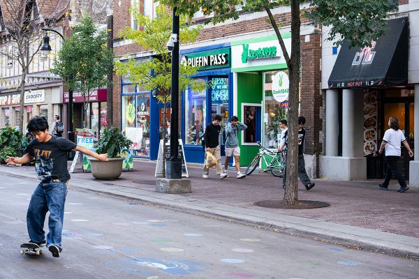 A skateboarder glides down a lively street lined with shops, trees, and colorful chalk markings on the pavement.