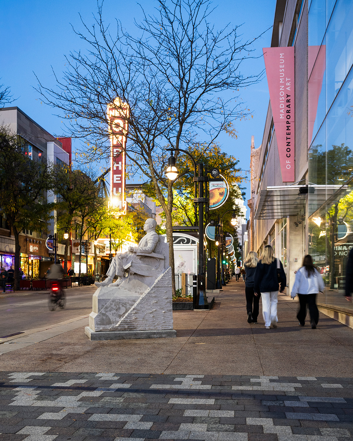 A statue sits along a pedestrian walkway near the Madison Museum of Contemporary Art as people stroll under streetlights at dusk. The striking Orpheum sign glows across the street.