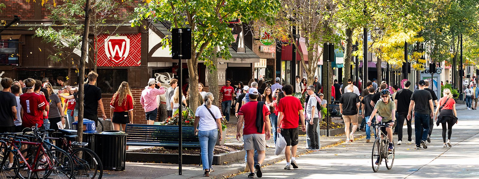A busy pedestrian street lined with trees and shops shows groups of people walking, biking, and gathering on a sunny day.