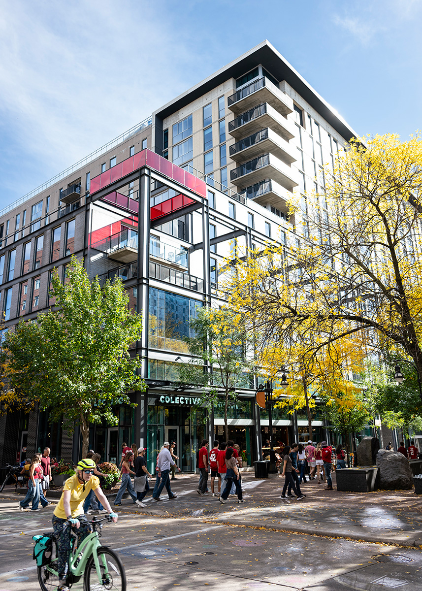 People walk and bike along a sunlit plaza in front of a modern multi-story building with glass balconies and autumn trees. Colectivo Coffee resides on the street-level floor.