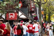 A crowd wearing red and white fills the sidewalk outside State Street Brats with festive signs and fall foliage overhead.