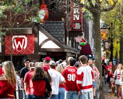 A crowd wearing red and white fills the sidewalk outside State Street Brats with festive signs and fall foliage overhead.