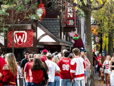 A crowd wearing red and white fills the sidewalk outside State Street Brats with festive signs and fall foliage overhead.