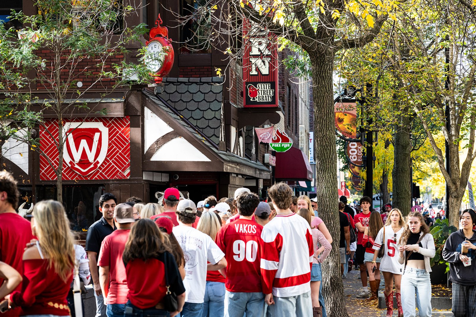 A crowd wearing red and white fills the sidewalk outside State Street Brats with festive signs and fall foliage overhead.