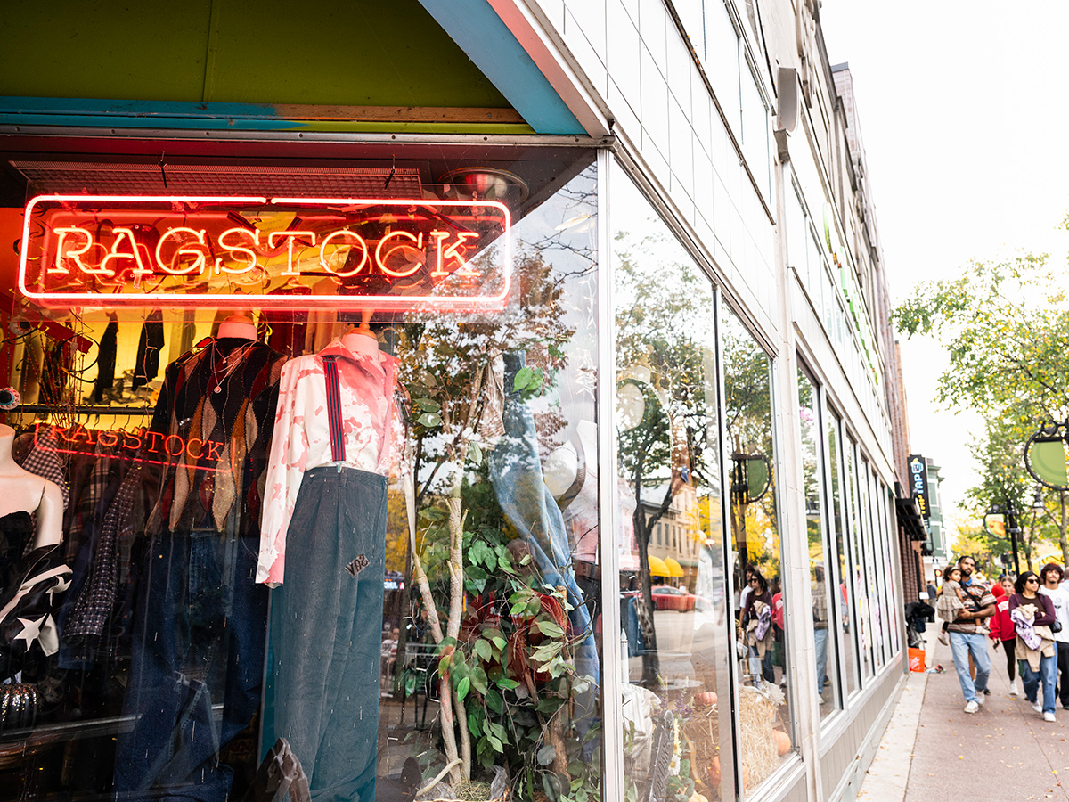 A storefront window displays mannequins wearing retro clothing beneath a bright red 'Ragstock' neon sign along a busy sidewalk.